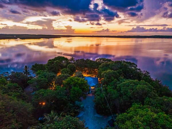 A tropical island at sunset with a blue-roofed house nestled among dense trees and a reflective water surface.