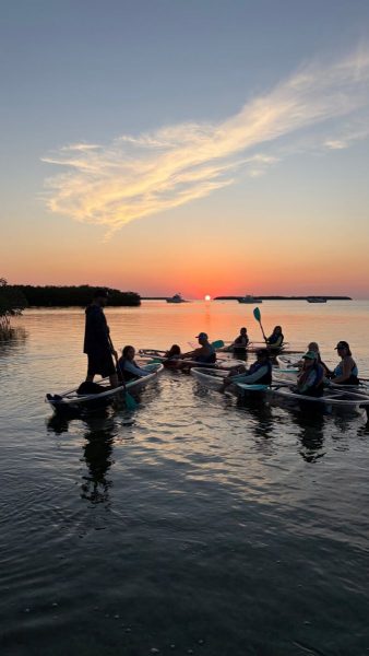 Group of people in kayaks around a guide on a calm coastal inlet at sunset, with warm skies reflected in the water.