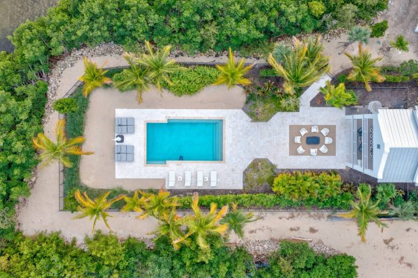 Aerial view of a rectangular pool surrounded by a stone patio, lounge chairs, and a dining area amid palm trees and tropical vegetation.