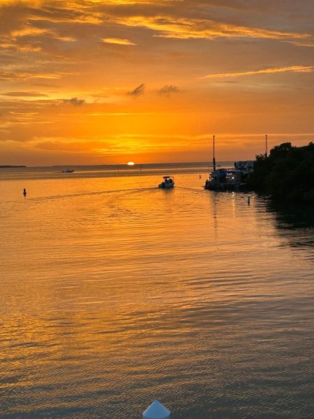 Golden sunset over a calm coastal inlet with orange sky, small boats on the water, and a docked marina on the right side.